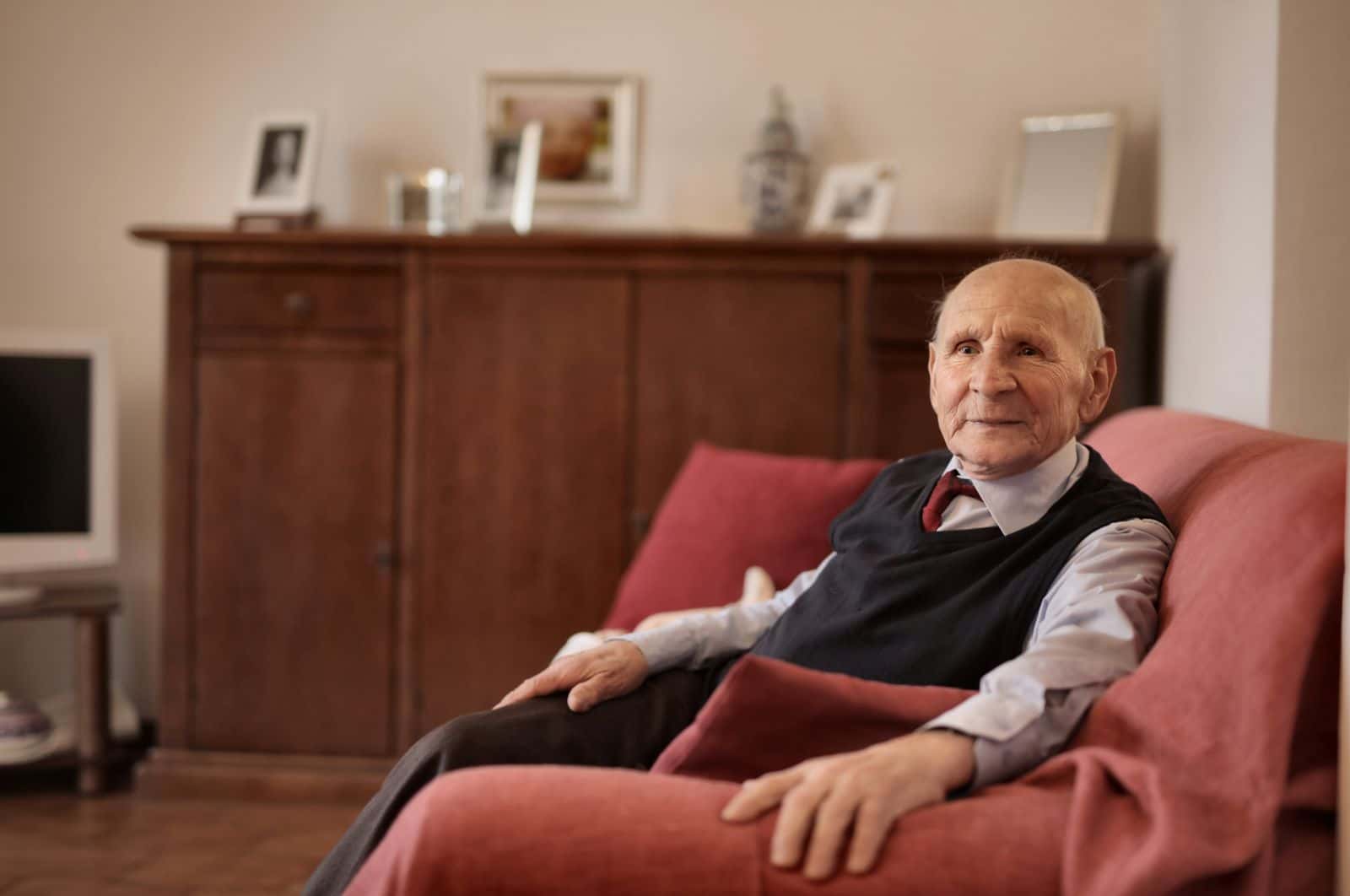Un homme âgé, assis dans un fauteuil rouge de son salon, regarde vers la fenêtre