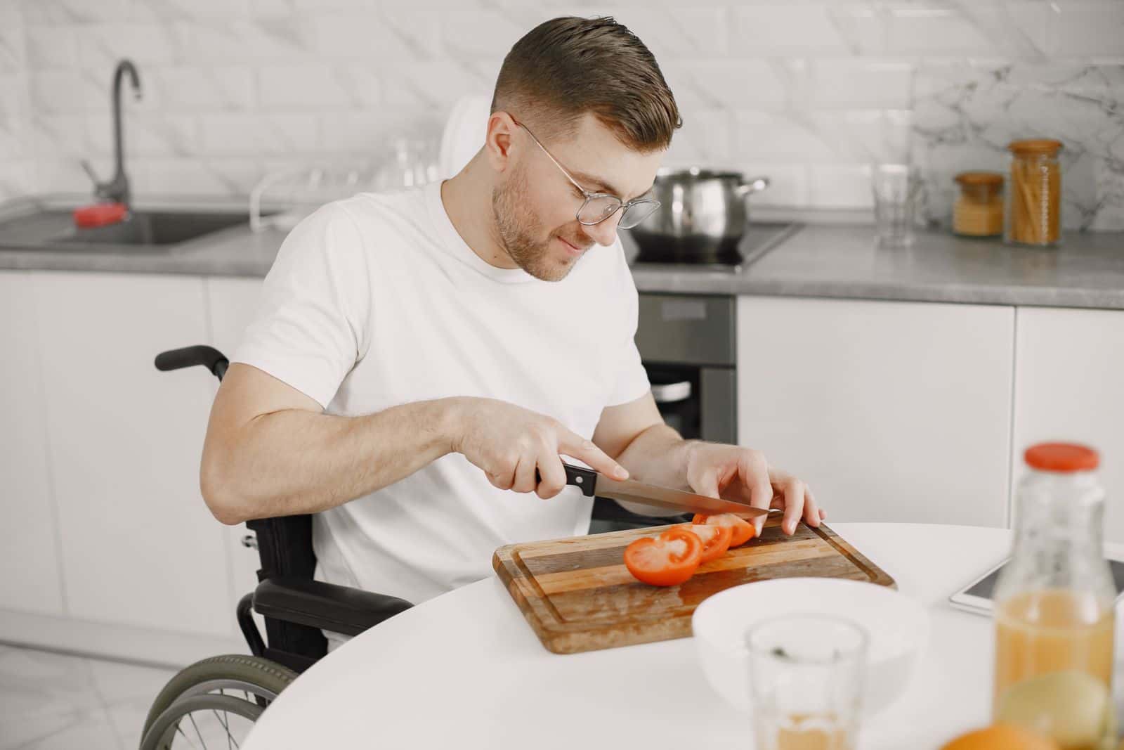 Un homme en fauteuil roulant coupe une tomate sur une planche dans une cuisine