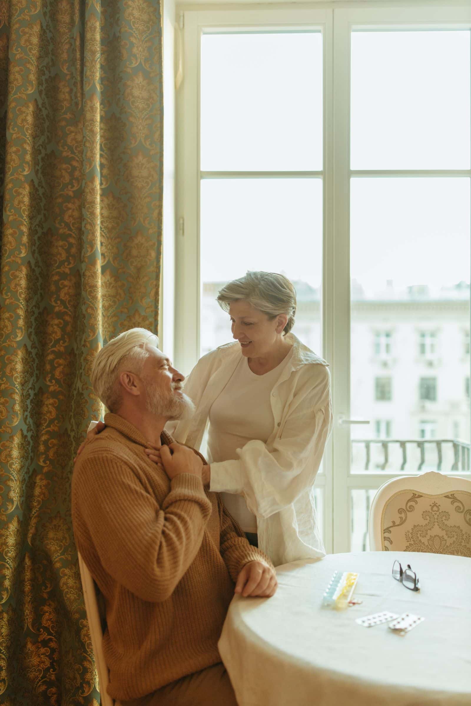 Une femme âgée debout pose la main sur l'épaule d'un homme âgé assis à une table, devant une fenêtre