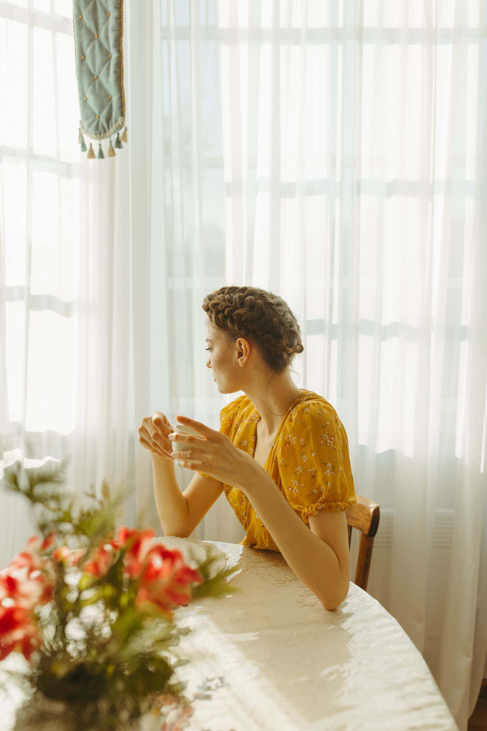 Une femme d'une trentaine d'années, assise à une table près d'une fenêtre, tient une tasse