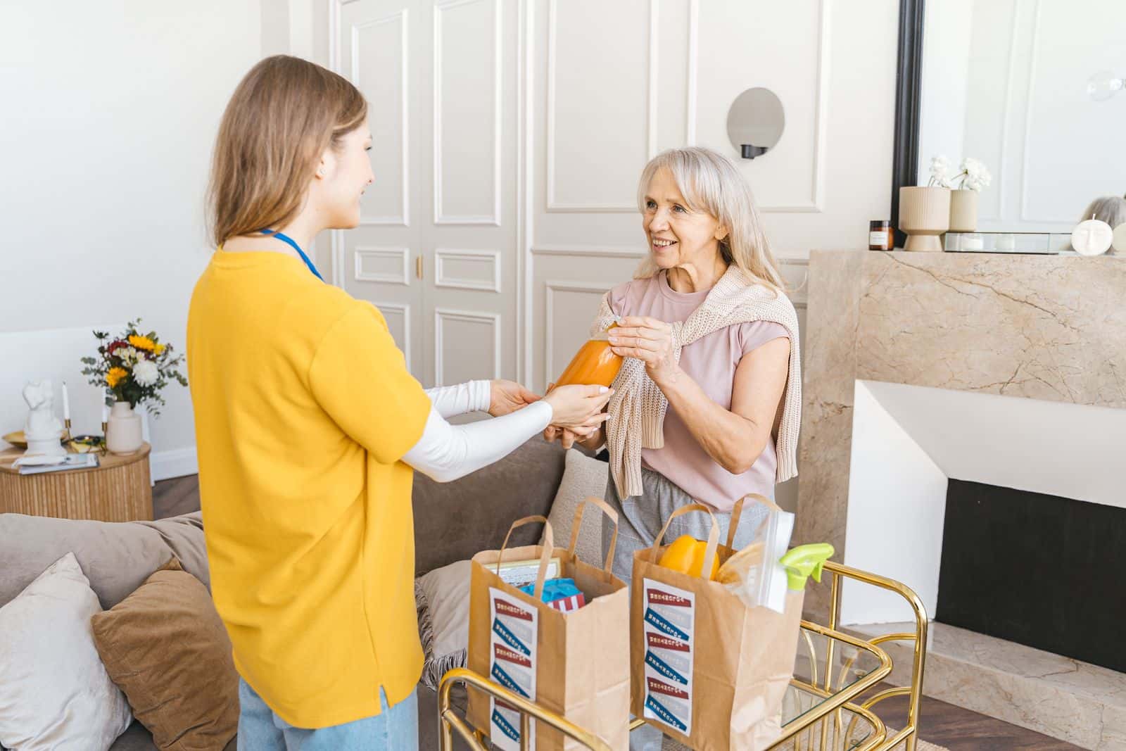 Une jeune femme tend un fruit à une femme âgée dans un salon, des sacs de courses posés au sol