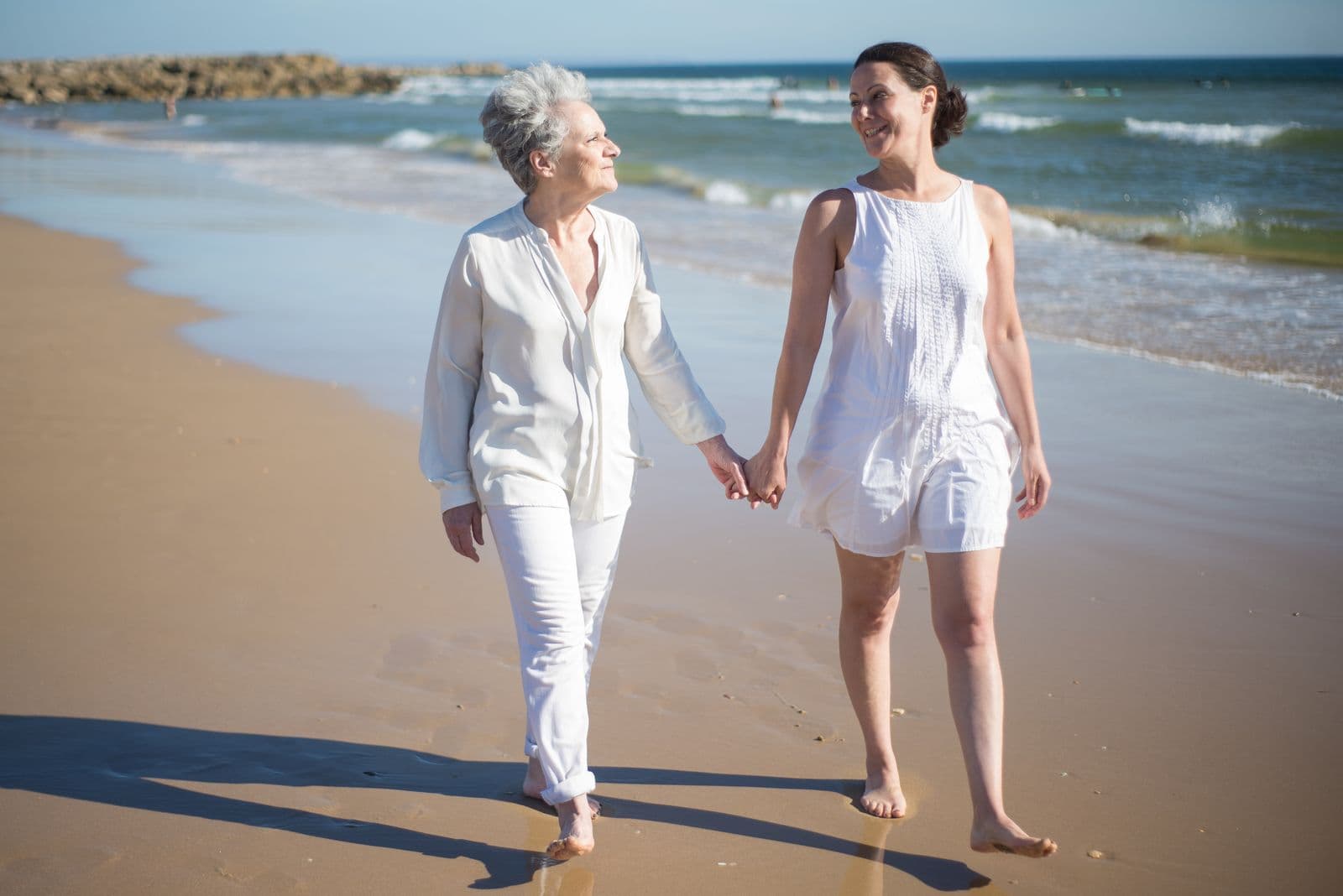 Une femme âgée et une femme plus jeune marchent main dans la main au bord de l'eau, sur une plage