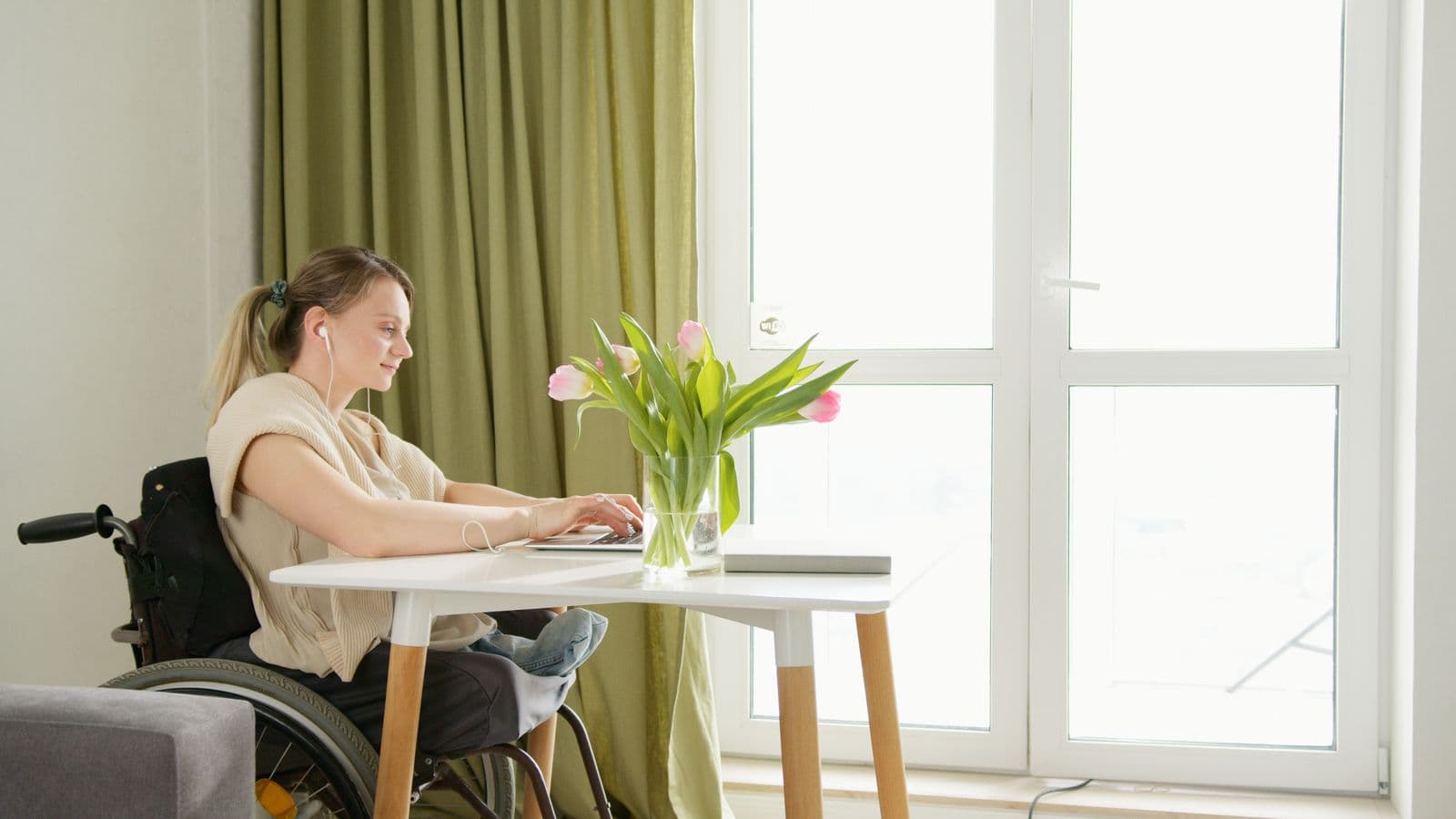 Une femme en fauteuil roulant travaille sur un ordinateur portable à une table, près d'une fenêtre, un bouquet de tulipes devant elle