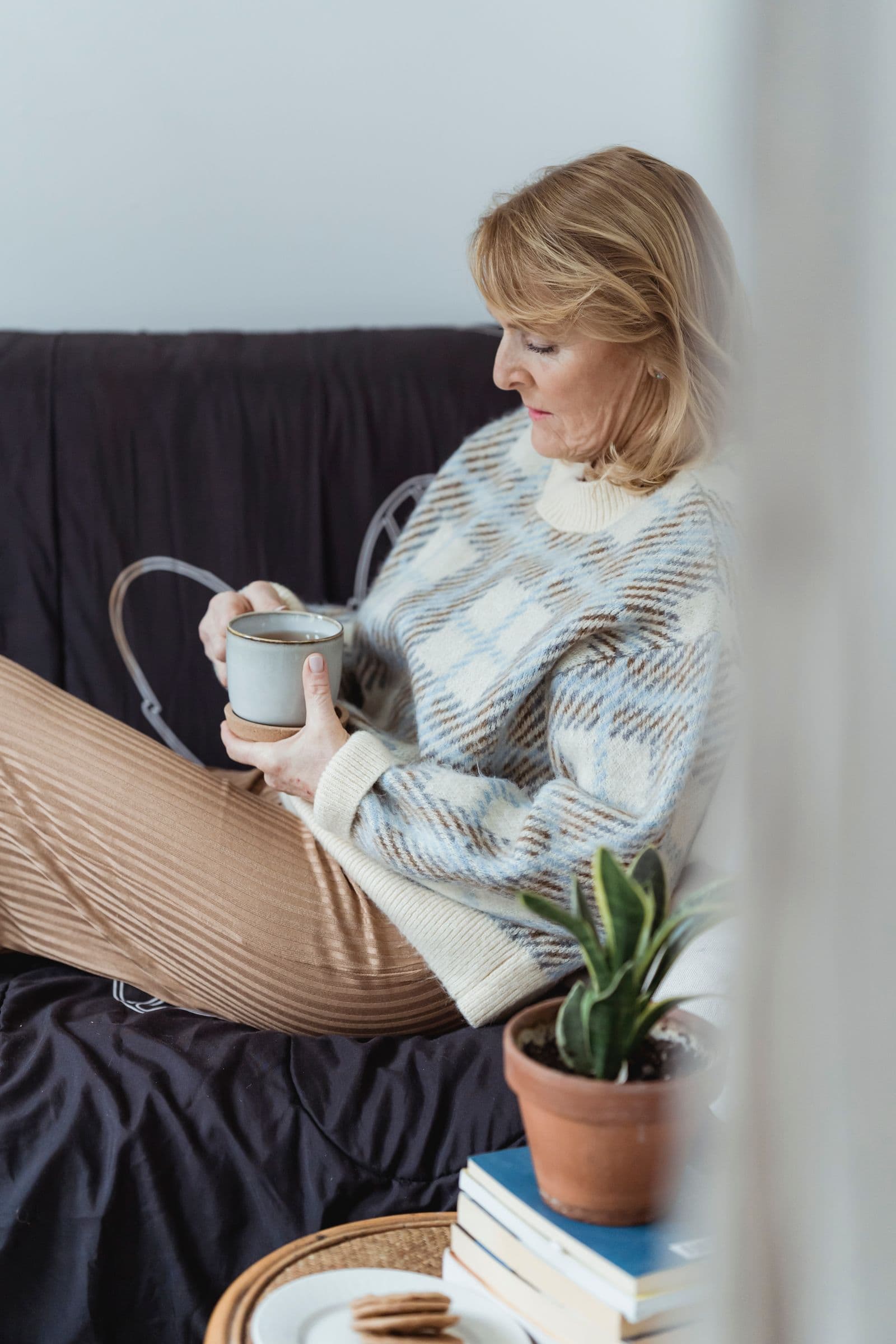 Une femme aux cheveux blonds, assise sur un canapé, tient une tasse et regarde vers le bas