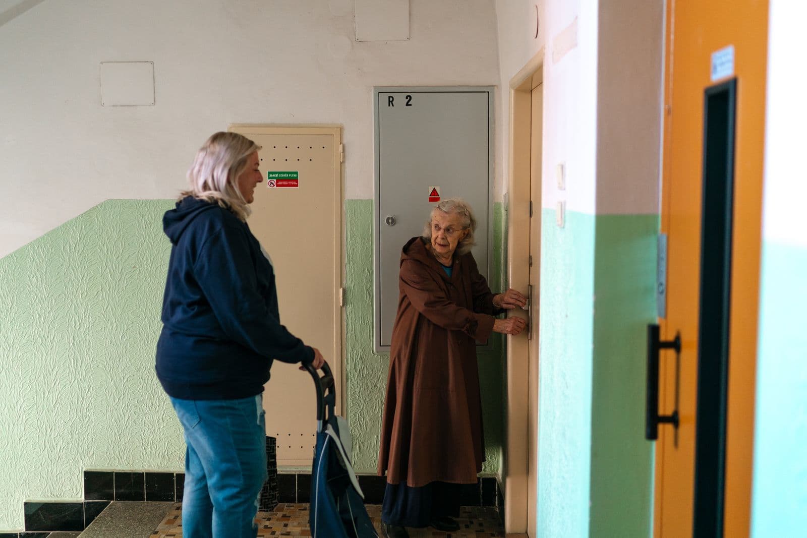 Sur un palier d'immeuble, une femme accompagne une femme âgée qui s'appuie sur un déambulateur devant sa porte