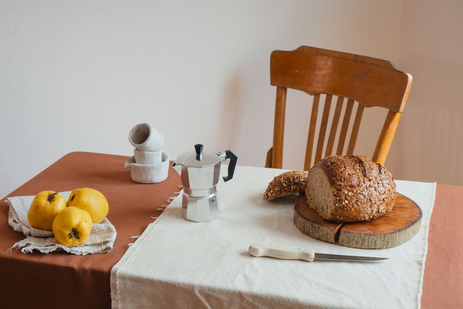 Une table de cuisine avec du pain, une cafetière italienne et des coings, sous une lumière douce