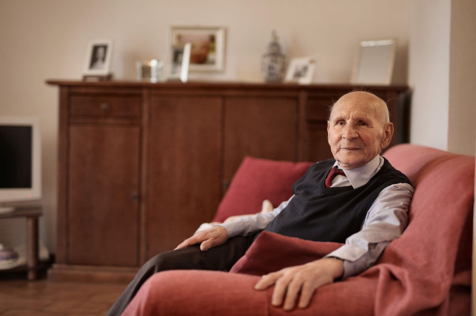 Un homme âgé, assis dans un fauteuil rouge de son salon, regarde vers la fenêtre