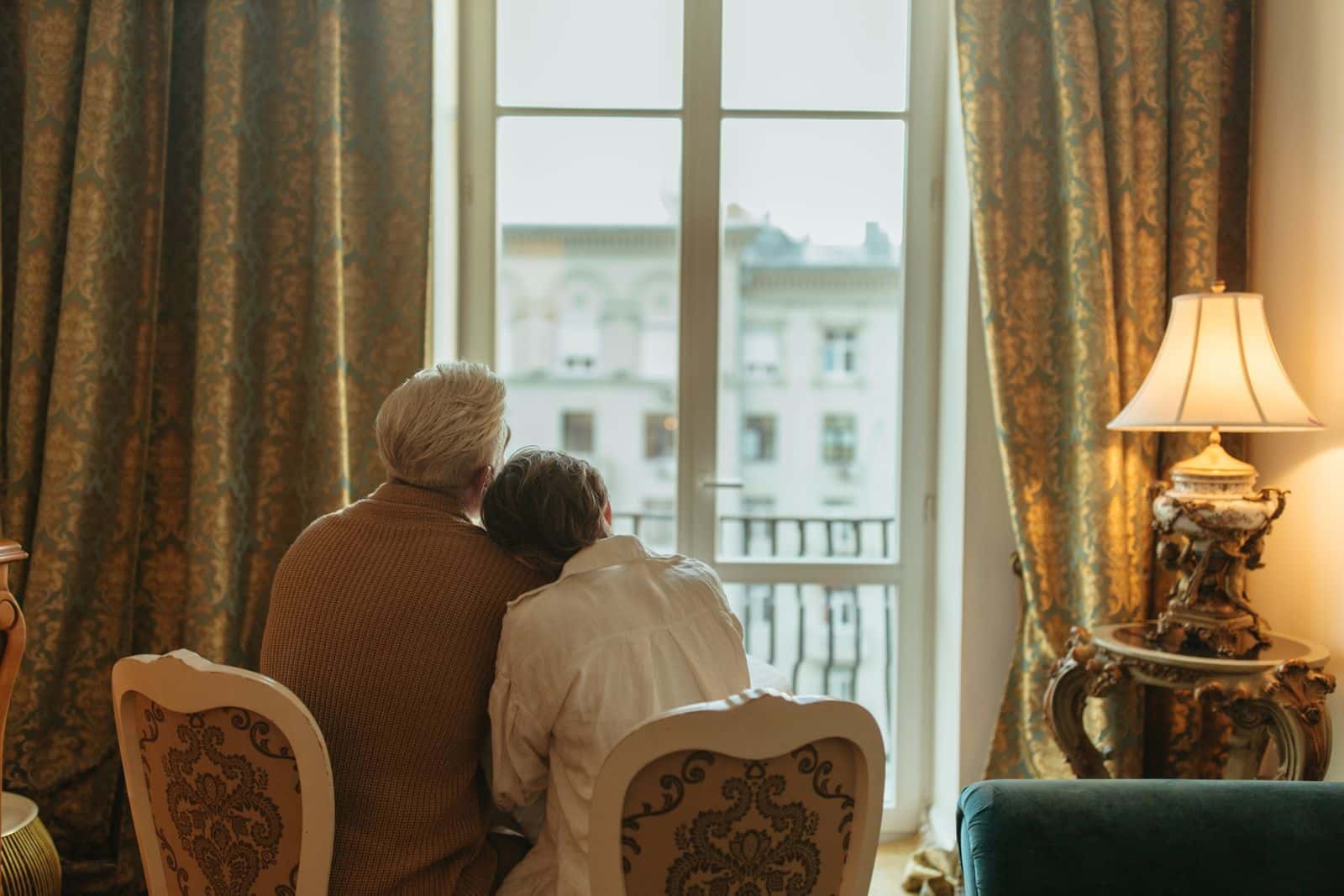Un couple âgé, vu de dos, assis côte à côte, regarde par une grande fenêtre