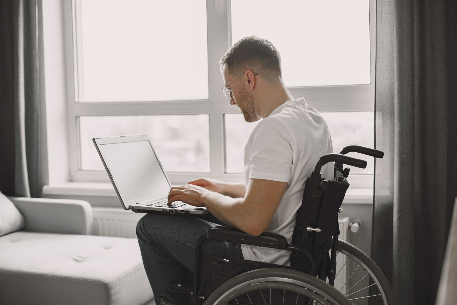 Un homme en fauteuil roulant travaille sur un ordinateur portable près d'une grande fenêtre
