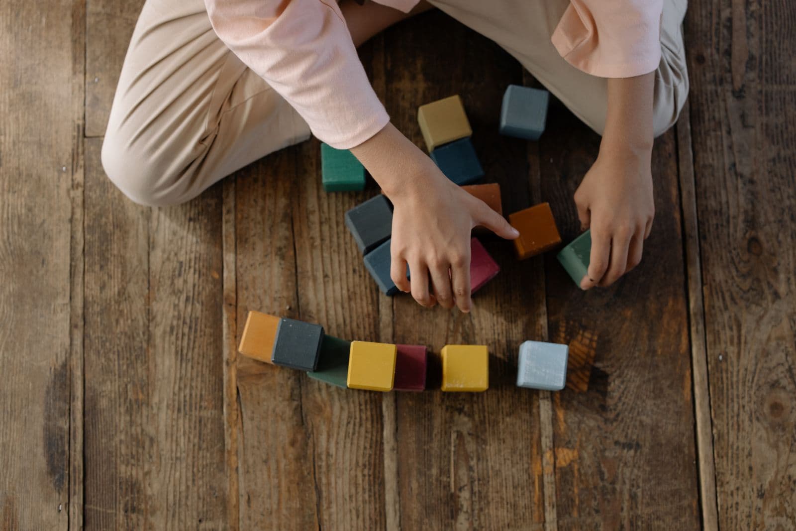 Les mains d'un enfant, vu de haut, alignent des cubes de bois colorés sur un plancher
