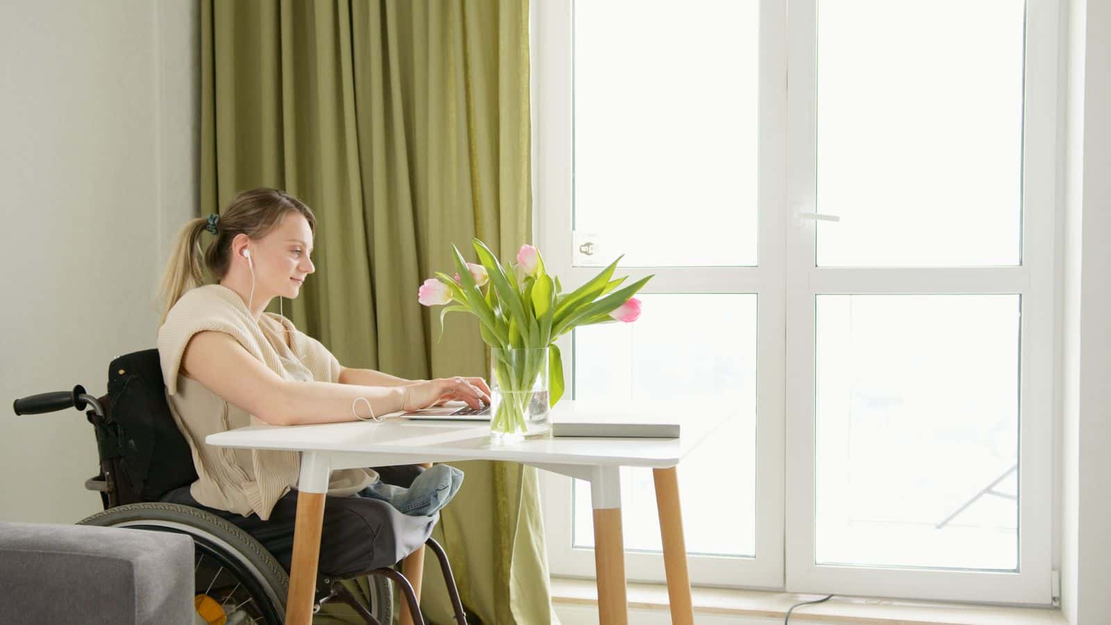 Une femme en fauteuil roulant travaille sur un ordinateur portable à une table, près d'une fenêtre, un bouquet de tulipes devant elle