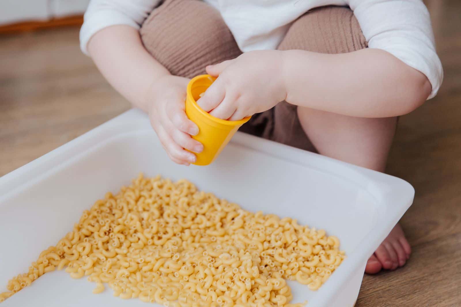 Un enfant, dont on ne voit que les mains et les jambes, joue avec des pâtes sèches dans un bac blanc