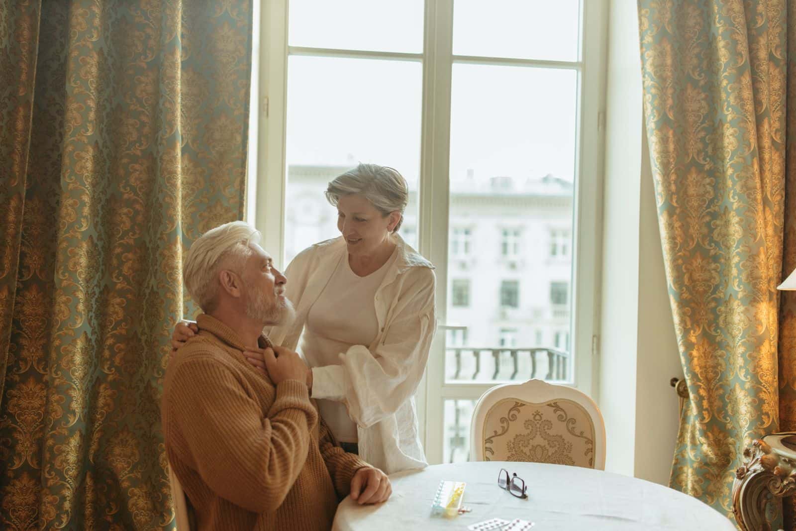 Une femme âgée debout parle avec un homme âgé assis à une table, dans une pièce baignée de lumière