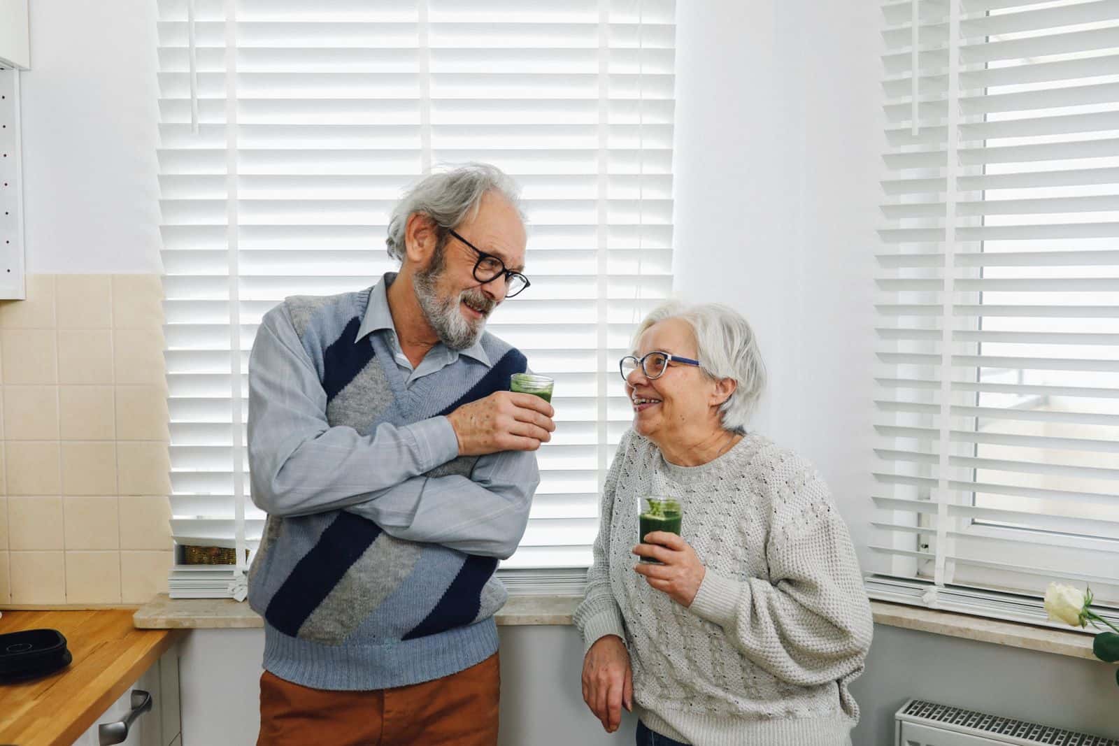 Un couple âgé, debout dans une cuisine, boit un verre devant la fenêtre