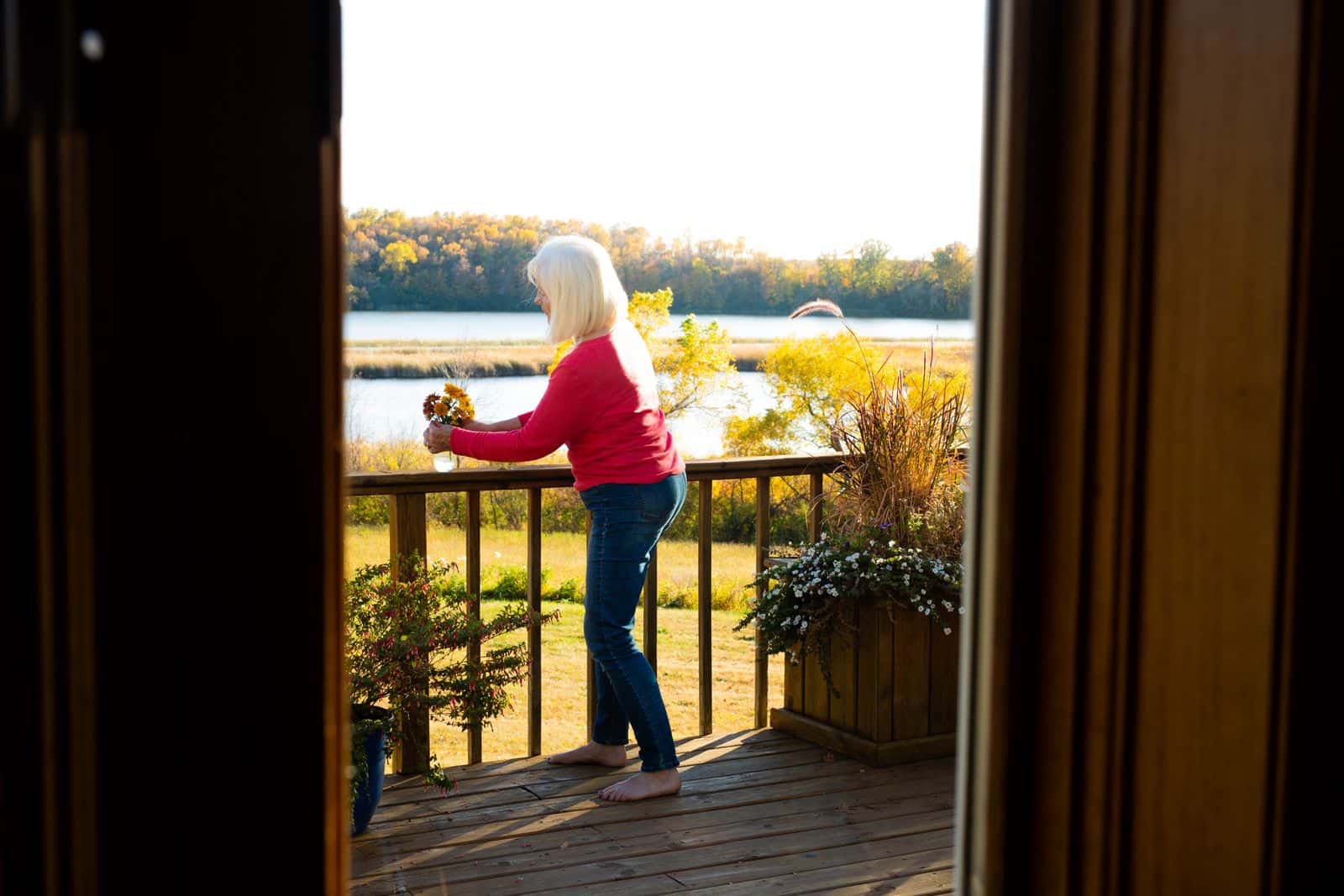Vue depuis l'intérieur : une femme aux cheveux blancs se tient sur une terrasse en bois face à un lac, au soleil